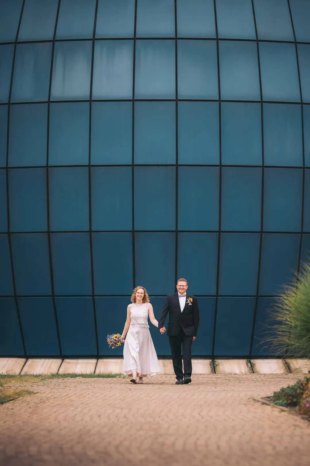 hochzeitsfotograf-wolfsburg-planetarium-kuppel Ein Brautpaar spaziert Hand in Hand mit strahlenden Gesichtern vor der Kuppel des Planetarium Wolfsburg