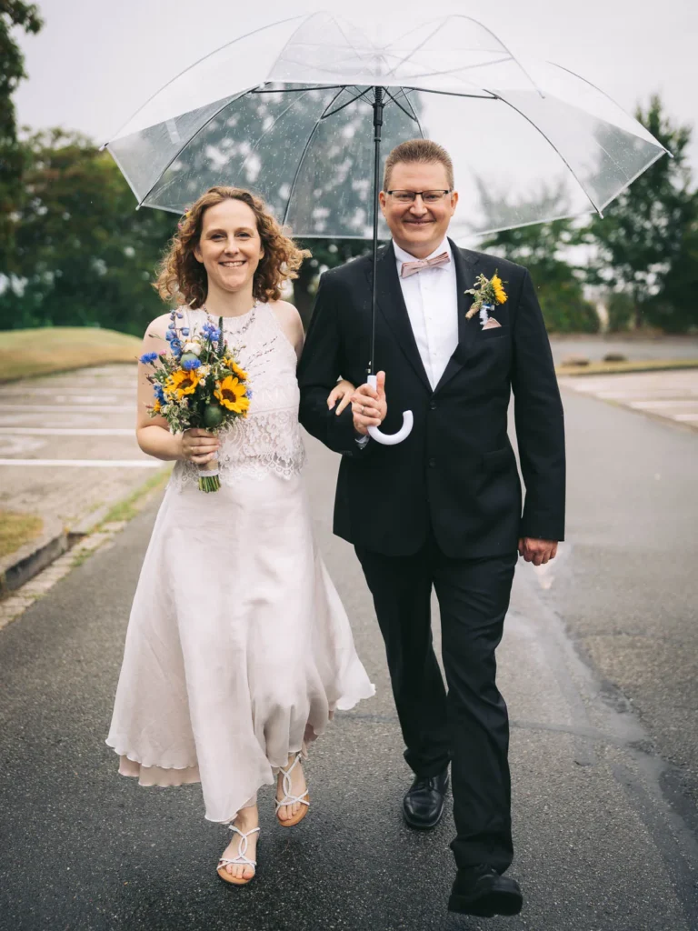 Ein Brautpaar spaziert an seinem regenreichen Hochzeitstag mit einem Regenschirm über den Parkplatz am Theater Wolfsburg.