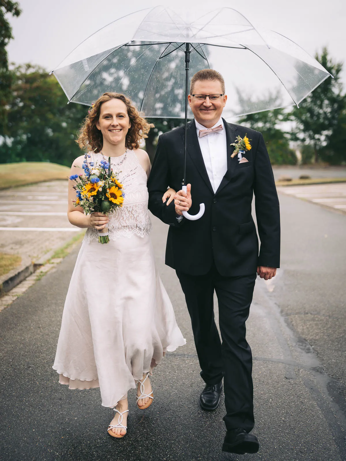 Ein Brautpaar spaziert an seinem regenreichen Hochzeitstag mit einem Regenschirm über den Parkplatz am Theater Wolfsburg.