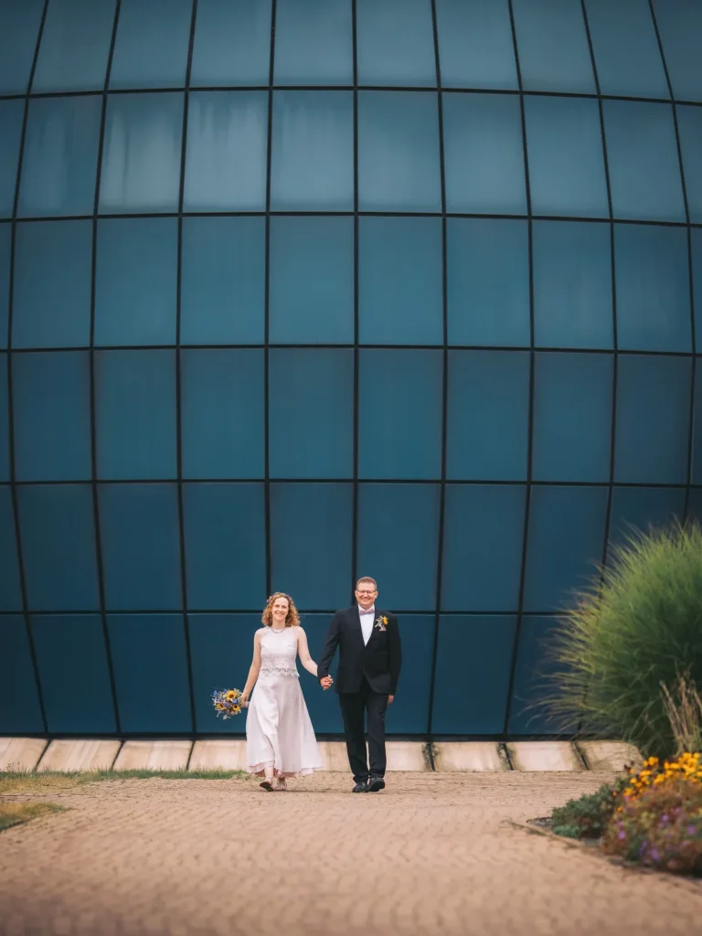 Ein glückliches frisch verheiratetes Ehepaar geht Hand in Hand auf vor dem Planetarium Wolfsburg auf den Hochzeitsfotografen zu