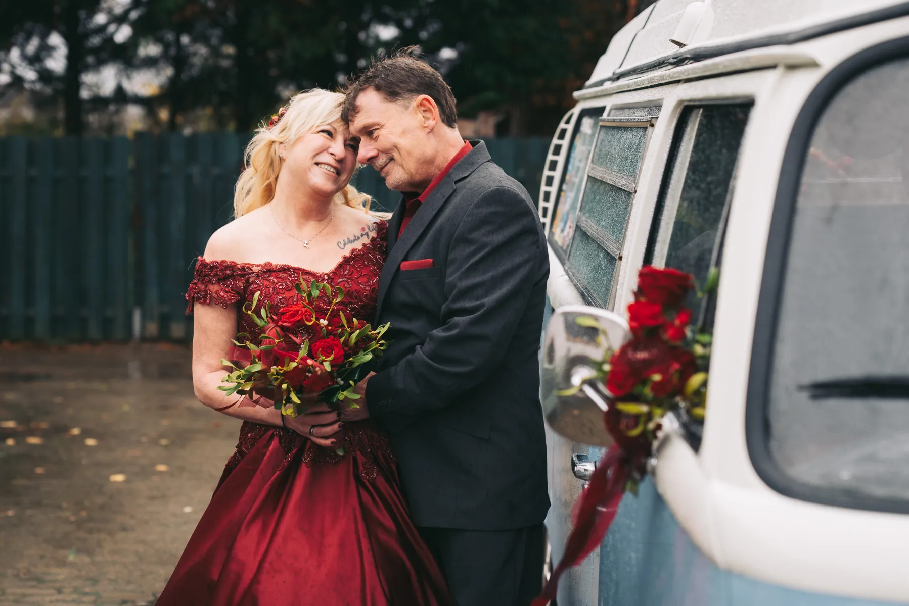 Ein verliebtes Brautpaar posiert nach der Hochzeit für ein Fotoshootings mit einem VW T1 Bus. Sie trägt ein rotes Kleid und er einen dunklen Anzug mit rotem Hemd. Der Brautstrauss und die Dekoration sind passend dazu auch in rot.