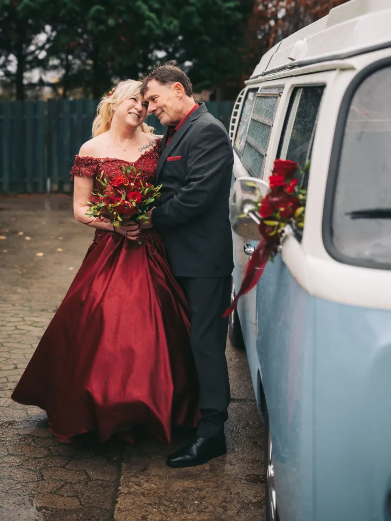 Ein Brautpaar posiert für eine Fotoshootings nach der Hochzeit mit einem alten VW Bus. Sie trägt ein rotes Brautkleid passend zum Brautstrauss und zur roten Blumendekoration.