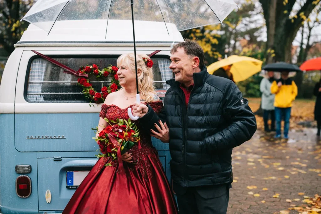 Brautpaar steht vor der Hochzeit im Schloss Fallersleben in Wolfsburg unter einem Regenschirm an einem VW Bus